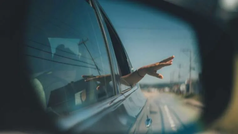 A passenger's hand out of a car window, seen in the wing mirror on a sunny road